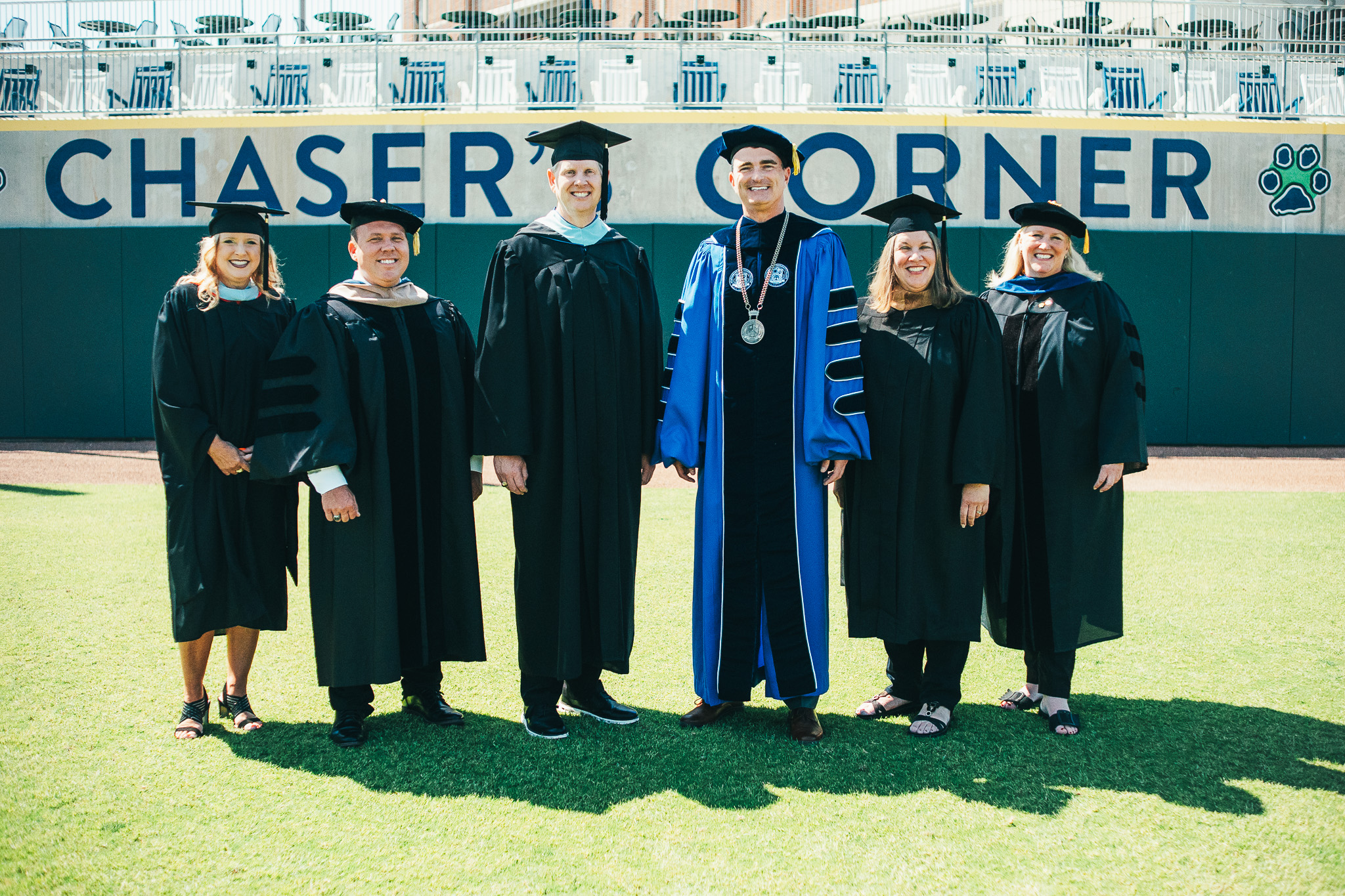 Six individuals wearing academic regalia stand on a grassy field in front of a stadium wall that reads