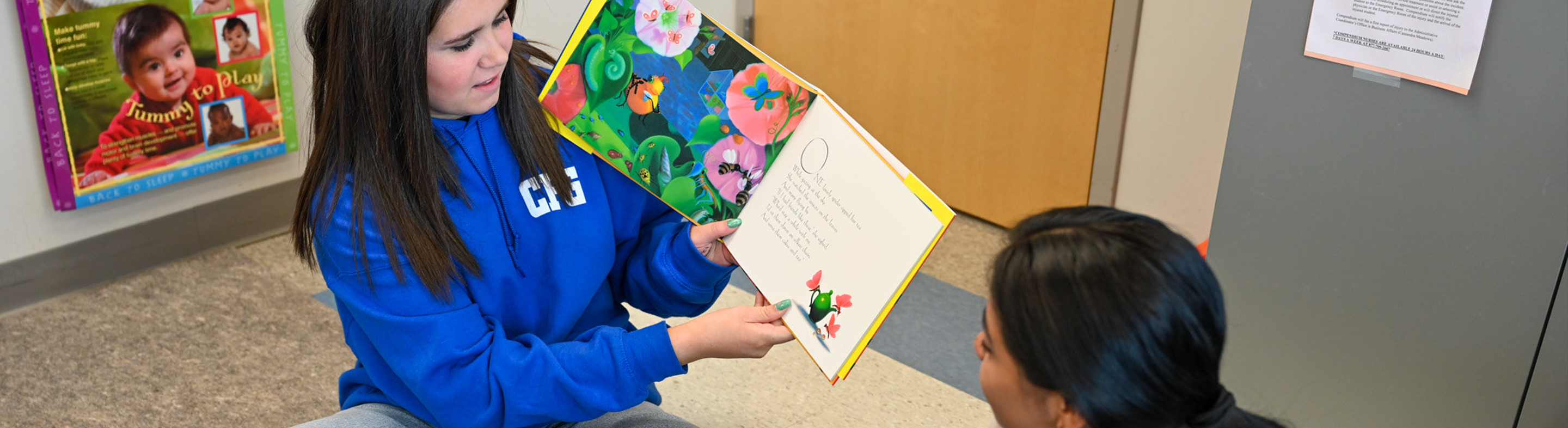 Image of student sitting on floor reading a children's book to someone.
